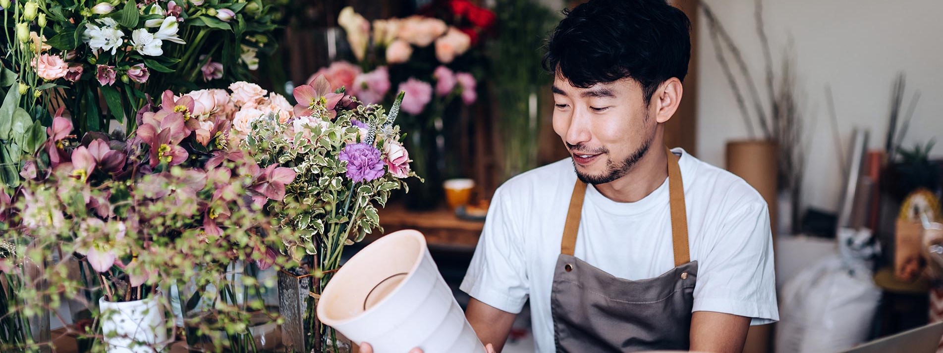 Young Asian Male Florist Working On Laptop Over Counter Against Flowers And Plants
